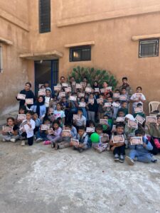 مجموعة كبيرة من الأطفال، أولاد وبنات، يجلسون في ساحة المدرسة ويحملون شهادات مشاركة في يوم كشفي.