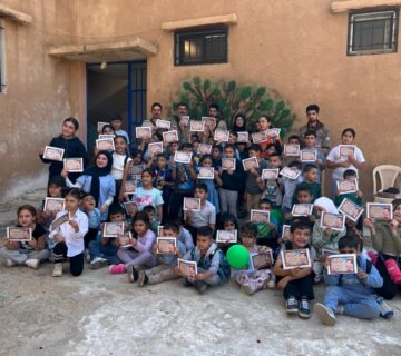 مجموعة كبيرة من الأطفال، أولاد وبنات، يجلسون في ساحة المدرسة ويحملون شهادات مشاركة في يوم كشفي.