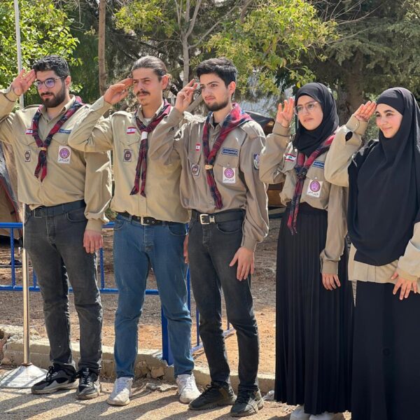 ثلاثة قادة كشفيين وقائدتان بزي الكشاف الرسمي يؤدون التحية الكشفية في الهواء الطلق.