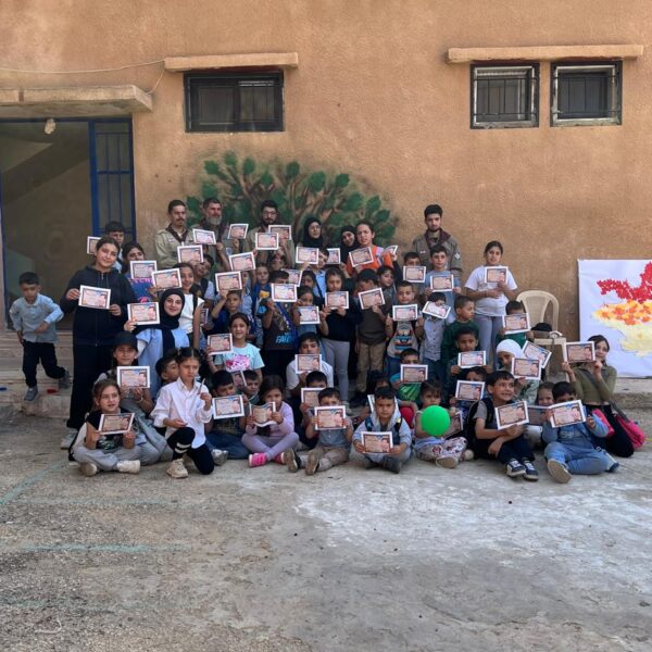 مجموعة كبيرة من الأطفال، أولاد وبنات، يجلسون في ساحة المدرسة ويحملون شهادات مشاركة في يوم كشفي.