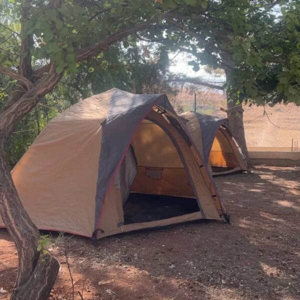 خيمة تخييم منصوبة تحت الأشجار وبجانبها موقد نار حجري في ساحة ترابية.