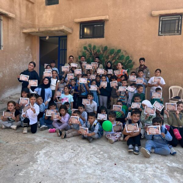 مجموعة كبيرة من الأطفال، أولاد وبنات، يجلسون في ساحة المدرسة ويحملون شهادات مشاركة في يوم كشفي.