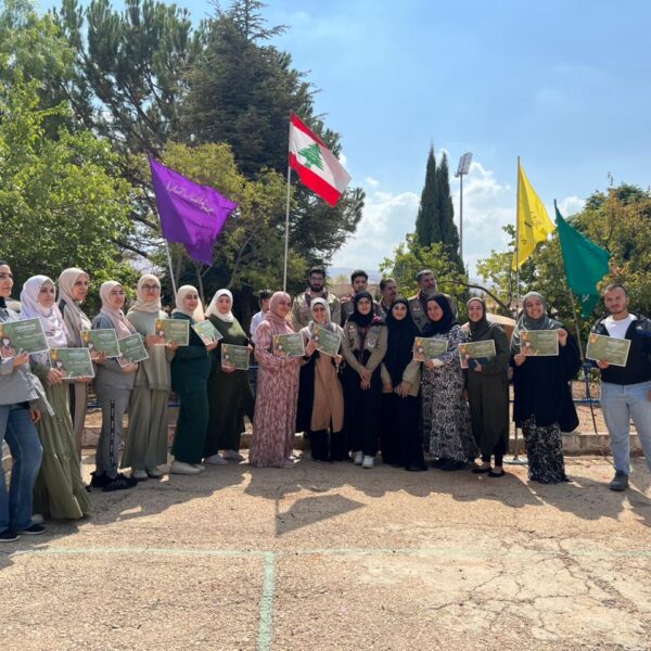 مجموعة من المعلمات يقفن بجانب قادة كشفيين ويحملن شهادات مشاركة أمام ساحة المدرسة.