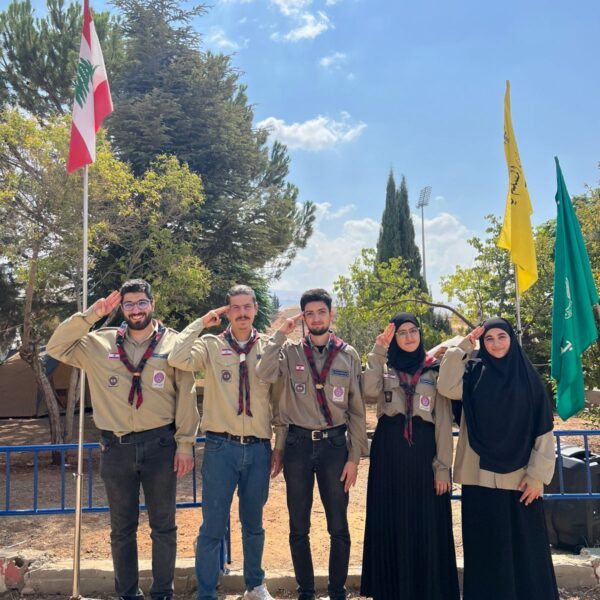 ثلاثة قادة كشفيين وقائدتان بزي الكشاف الرسمي يؤدون التحية الكشفية في الهواء الطلق.